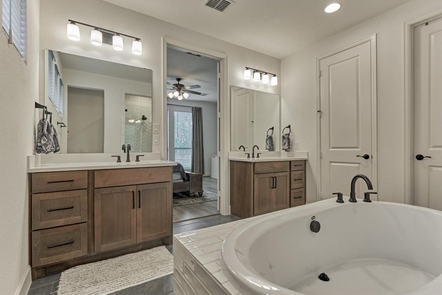 This photo showcases a modern bathroom featuring dual sinks with wooden cabinets, a large bathtub, and stylish fixtures. The room is well-lit with contemporary lighting and has a view into an adjoining bedroom.