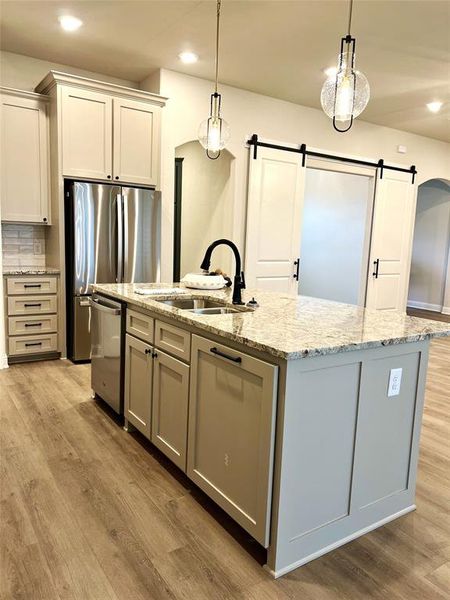 Kitchen with stainless steel appliances, a sink, a barn door, an island with sink, and light wood-type flooring Kitchen with stainless steel appliances, a sink, a barn door, an island with sink, and light wood-type flooring