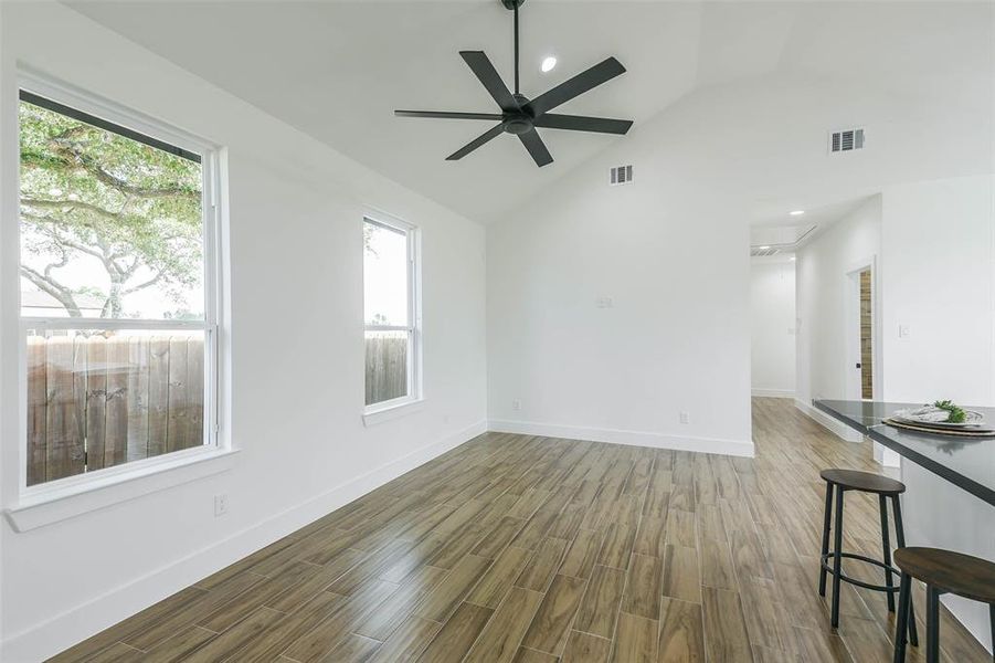 This is a bright, open-concept room with high ceilings, contemporary wood flooring, large windows for natural light, and a modern ceiling fan. The space is versatile, with a view into another room and a peek at the kitchen bar area.