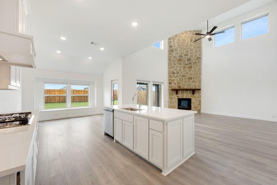 Open-concept kitchen with white island, light wood floors, and tall stone fireplace in living area Open-concept kitchen with white island, light wood floors, and tall stone fireplace in living area