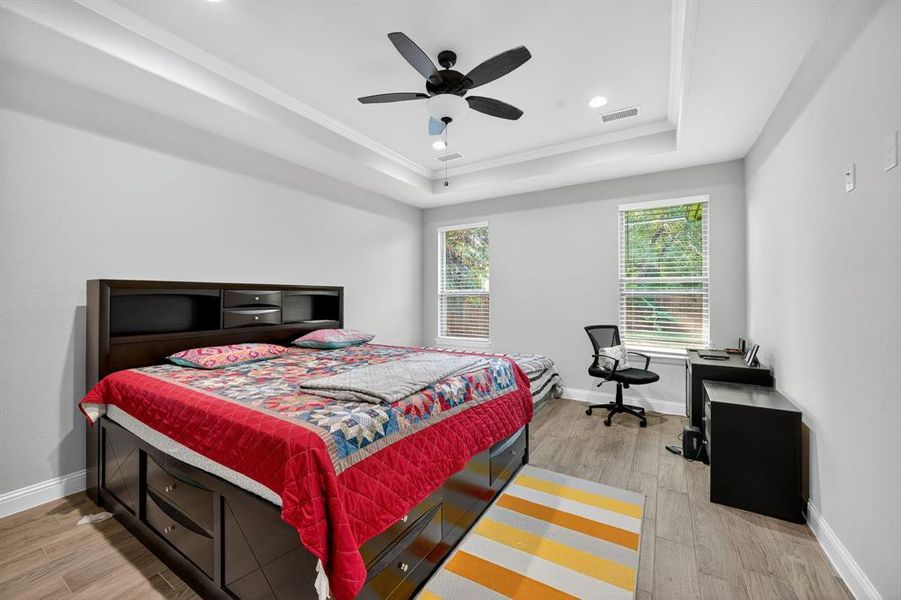 Bedroom featuring light wood-style floors, a raised ceiling, a ceiling fan, ornamental molding, and a desk