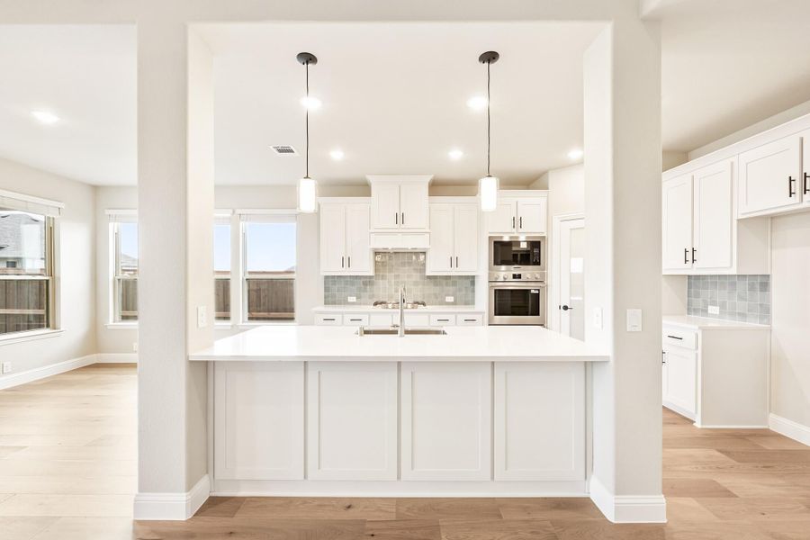 Kitchen with white cabinets, large island, pendant lights, tile backsplash, and stainless steel appliances on light hardwood floors