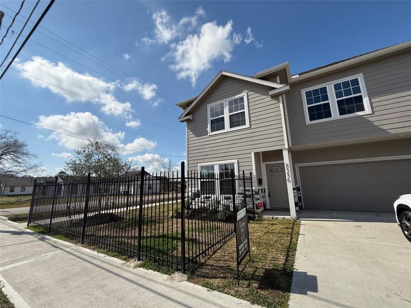 This photo shows a modern two-story home with a gray exterior, fenced front yard, and attached two-car garage. It is situated on a corner lot with a concrete driveway and sidewalk, under a clear, blue sky.