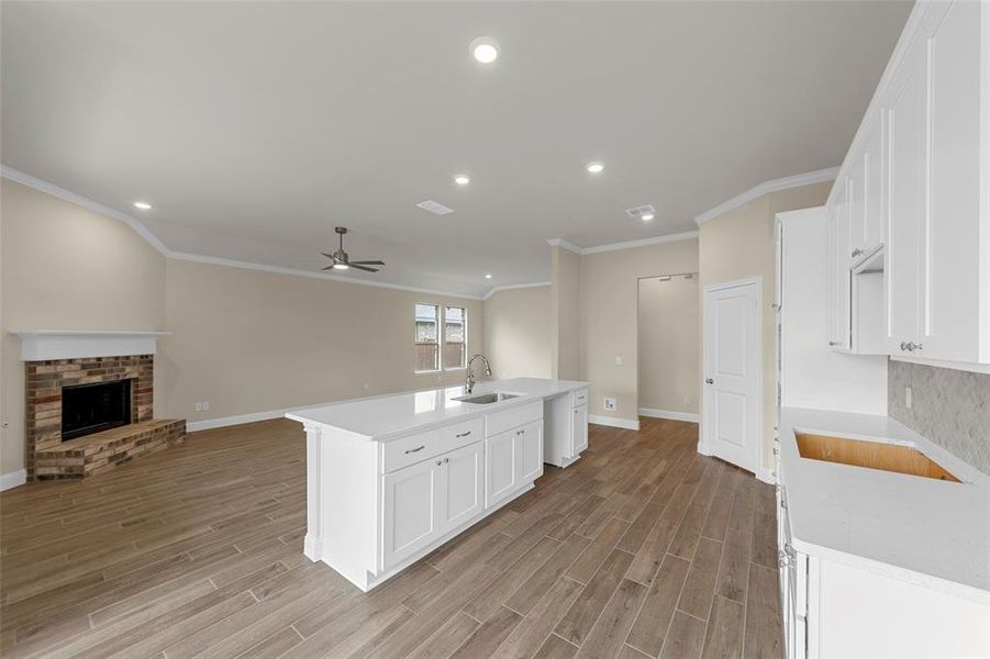 Open-concept living area featuring wood-finish flooring, a kitchen island with integrated sink, white cabinetry with stone countertops, a brick fireplace, and recessed lighting