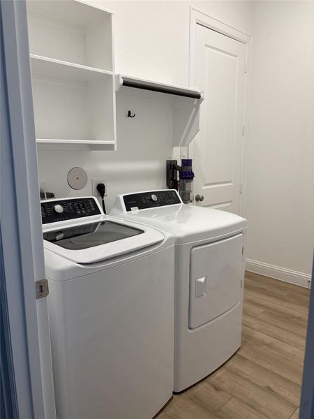 This is a modern laundry room featuring a washer and dryer set, white shelving, and a wood floor. It's well-organized with a clean, bright atmosphere.