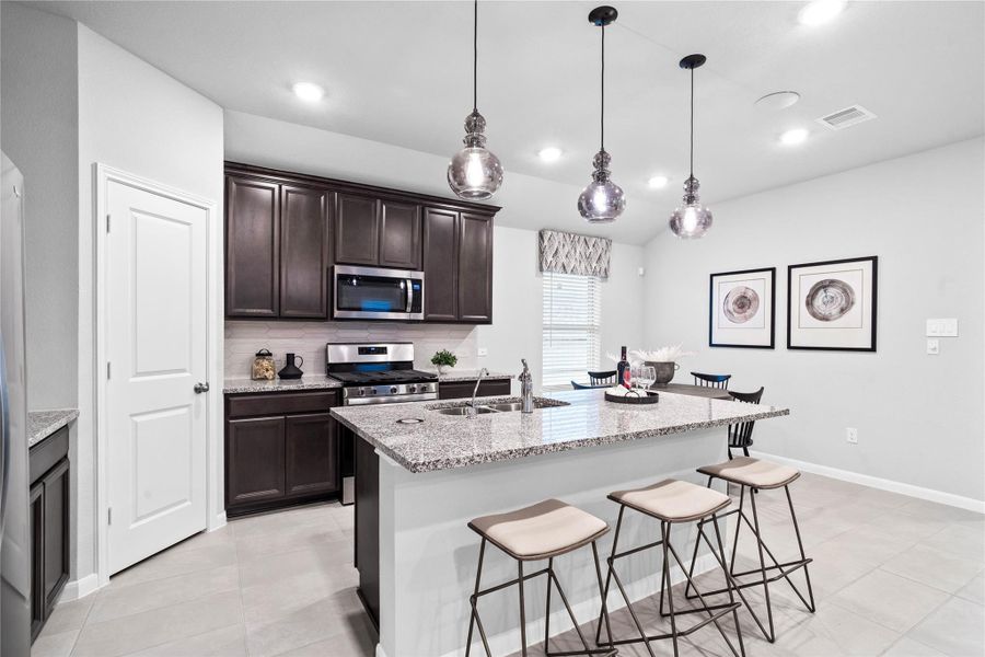 The expansive central island features a durable granite countertop, integrated sink, and an inviting breakfast bar with additional seating, illuminated by three elegant glass pendant lights.