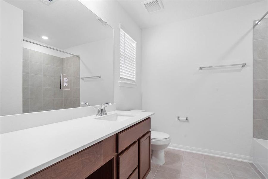 Secondary bath features tile flooring, bath/shower combo with tile surround, stained wood cabinets, beautiful light countertops, mirror, dark, sleek fixtures and modern finishes.