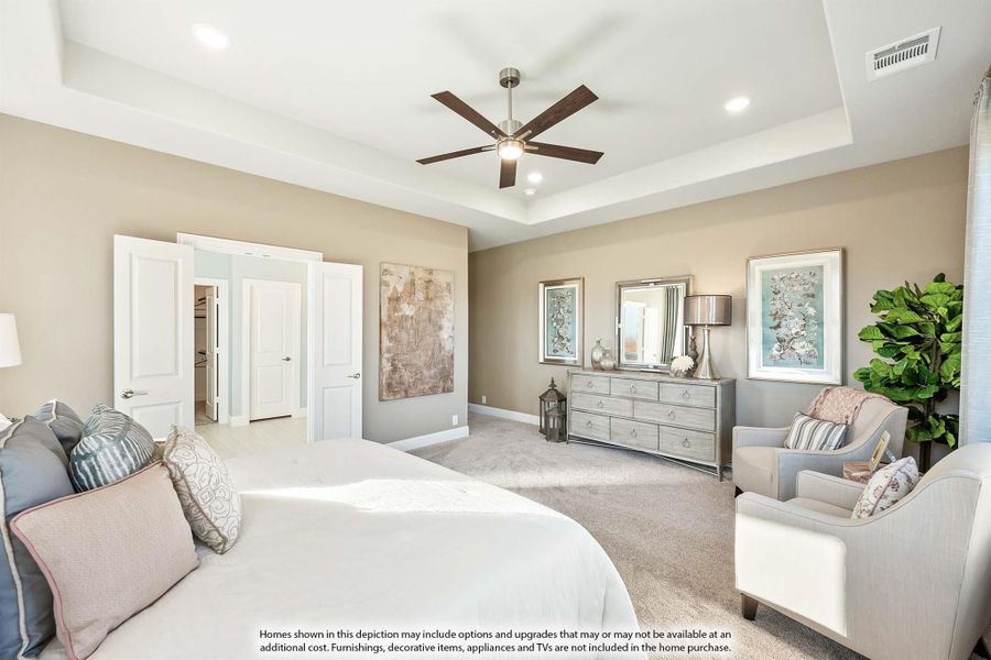 Master bedroom with tray ceiling, ceiling fan, carpet flooring, dresser, and seating area near walk-in closet entrance