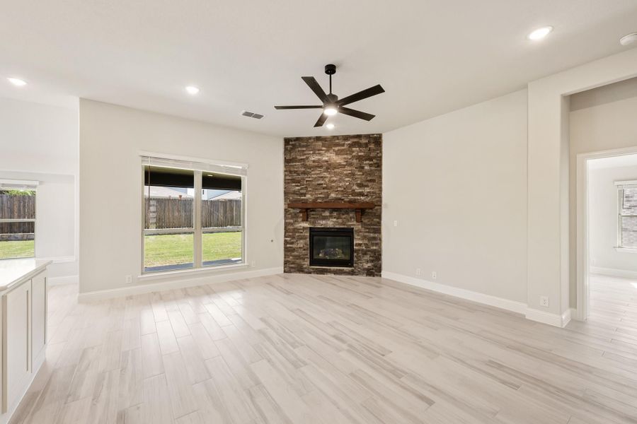 Open living room with stone fireplace, ceiling fan, light wood floors, and backyard window view Open living room with stone fireplace, ceiling fan, light wood floors, and backyard window view