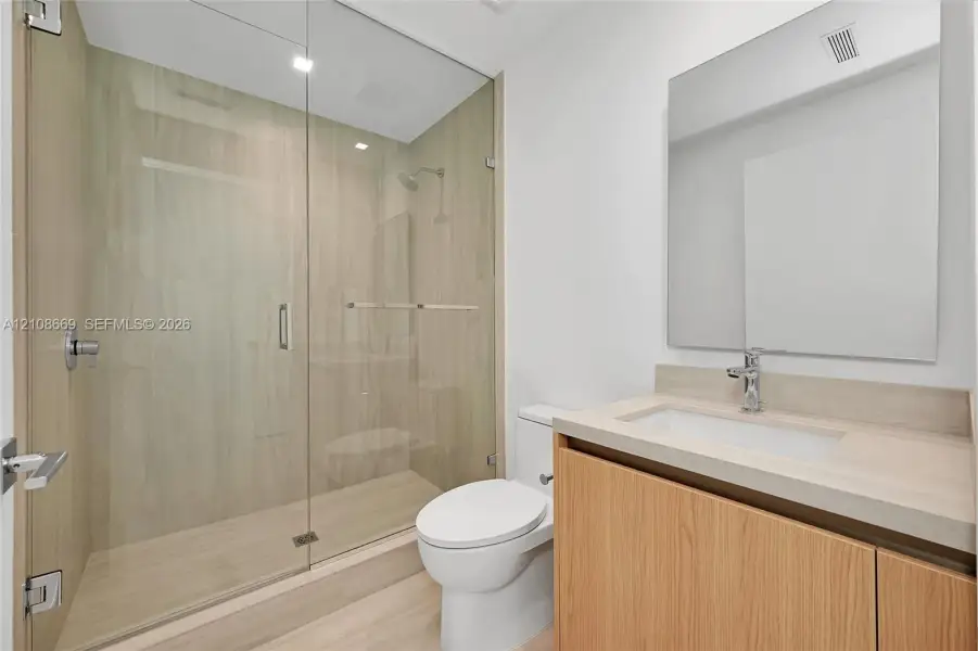 Full-height glass shower enclosure with integrated light-toned tile, wood-finish cabinetry, countertop-integrated basin, and frameless wall mirror