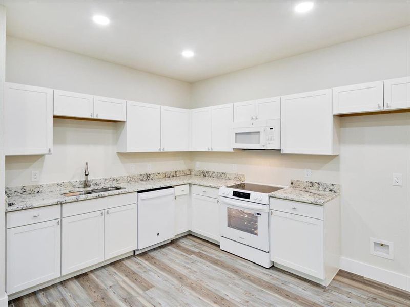 Bright kitchen featuring white cabinetry, granite-style countertops, wood-finish flooring, recessed lighting, and a full suite of white appliances including an oven and microwave Bright kitchen featuring white cabinetry, granite-style countertops, wood-finish flooring, recessed lighting, and a full suite of white appliances including an oven and microwave