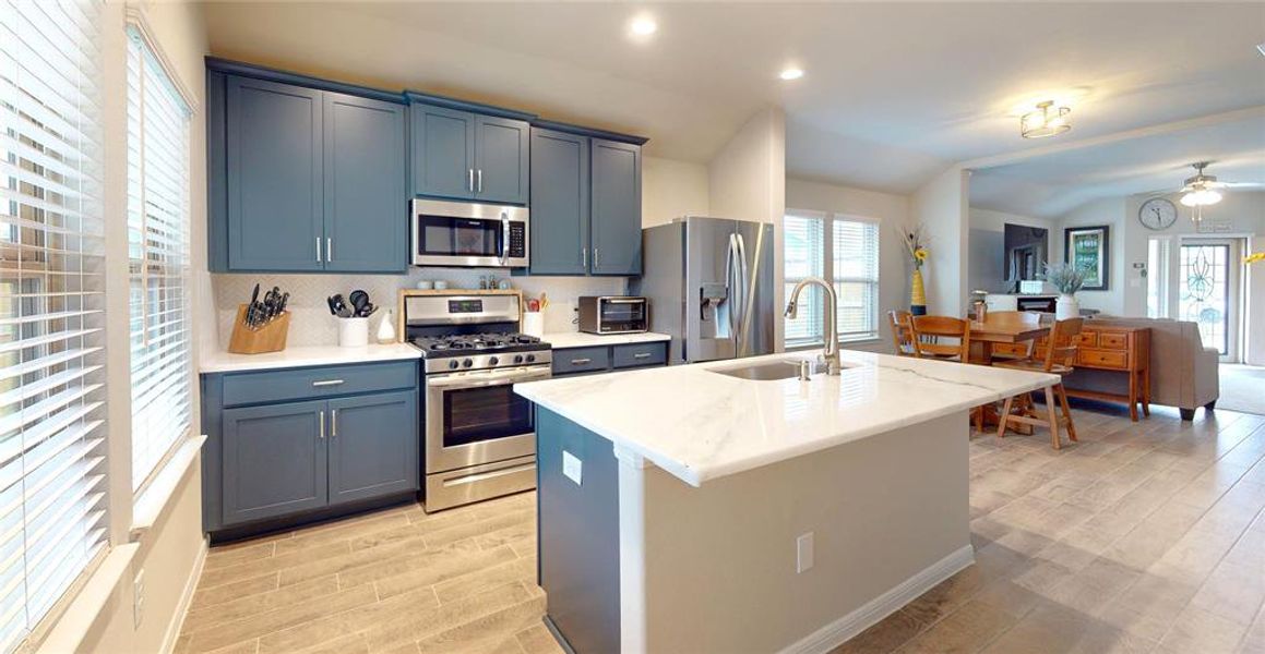 The kitchen features a stylish island with a sink and stainless steel fixtures set in a beautiful quartz countertop. Includes an upgraded dishwasher. The dining area is adjacent, leading to a cozy living space with a ceiling fan and large windows for natural light.  Note the overhang at the countertop that can accommodate breakfast stools too.