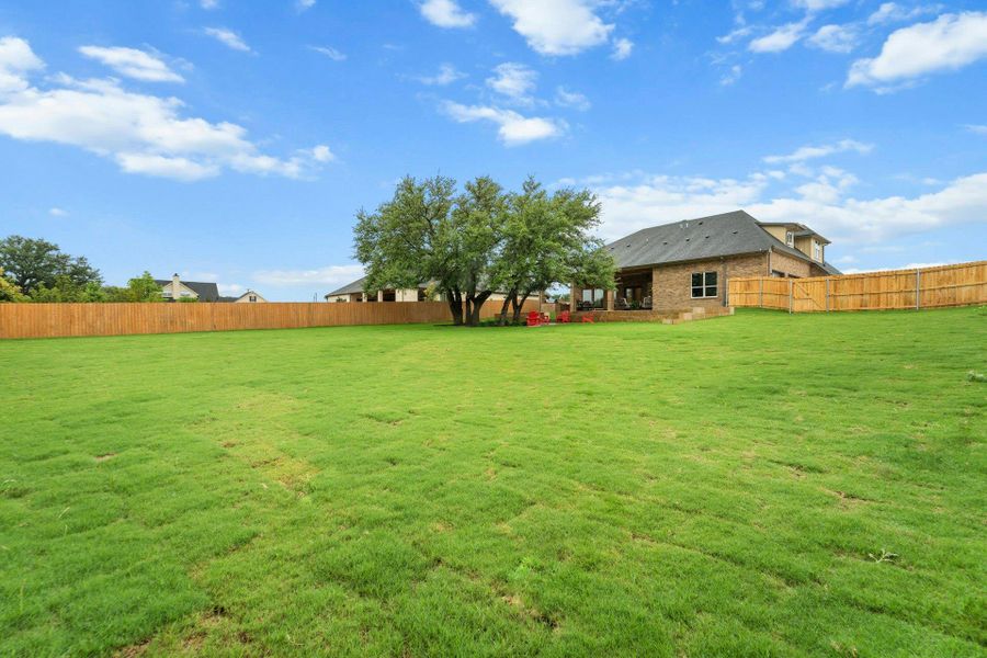Expansive grassy backyard featuring a full perimeter wood privacy fence, mature trees, and a brick-clad exterior with a covered patio