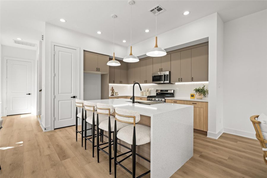 The kitchen features a large central island with a white-and-gray quartz waterfall countertop. It uses a clean two-tone design, pairing muted taupe upper cabinets with natural wood lower cabinets, all finished with simple black hardware and a sleek gooseneck faucet.