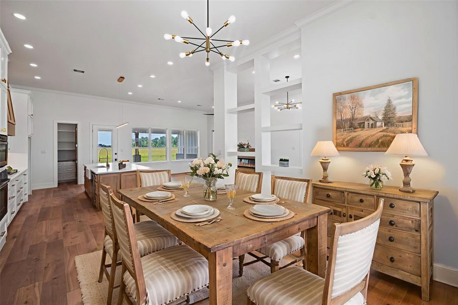 This spacious, open-concept kitchen and dining area features elegant lighting, a rustic wooden dining table, and a large island with a sink. The room is bright with natural light from large windows, offering a view of the outdoors. The decor is warm and inviting, with LVP floors and tasteful artwork. DIGITALLY STAGED