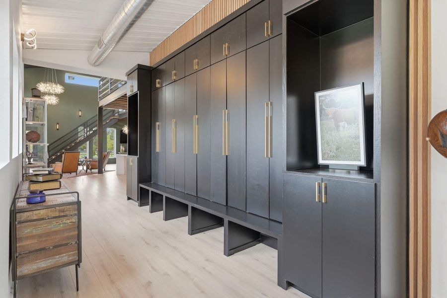 This photo showcases a modern interior with sleek black cabinetry and ample storage space. The room features light wood flooring, an industrial-style exposed duct, and a view into a bright living area with large windows and contemporary lighting fixtures.
