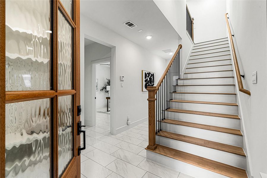 A bright entryway with a wooden front door, leading to a staircase with wooden steps and a metal railing. The space features white walls, tiled floors, and modern decor, creating a welcoming and contemporary atmosphere.
