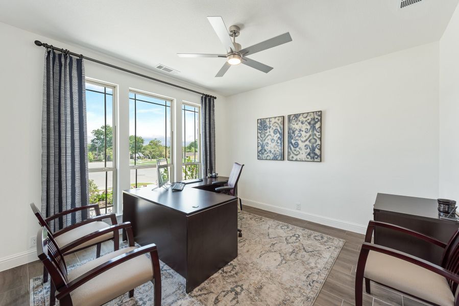 Home office with dark wood desk, ceiling fan, large windows, and area rug on tile floors