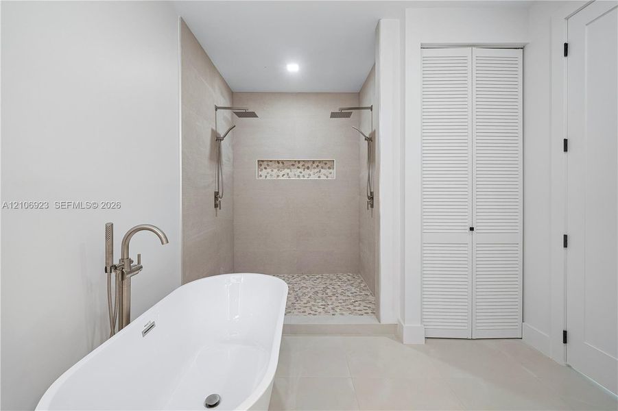 Freestanding soaking tub with metallic floor-mounted faucet, walk-in shower featuring dual overhead rainfall showerheads, recessed decorative tile niche, pebble-stone shower flooring, and louvered double-door cabinetry Freestanding soaking tub with metallic floor-mounted faucet, walk-in shower featuring dual overhead rainfall showerheads, recessed decorative tile niche, pebble-stone shower flooring, and louvered double-door cabinetry