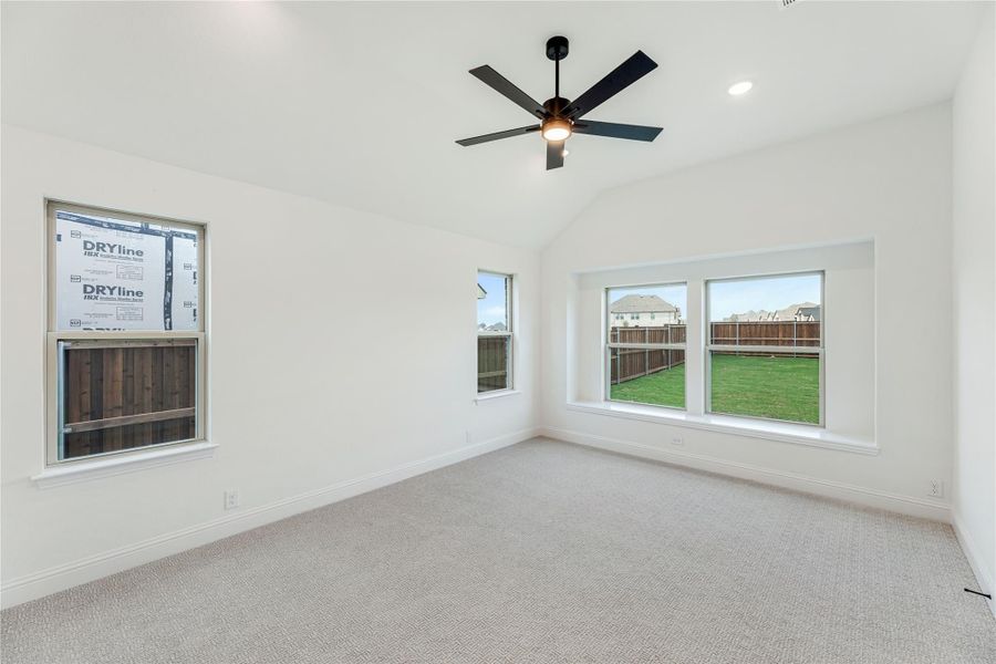 Empty bedroom with vaulted ceiling, black ceiling fan, carpet flooring, and multiple windows overlooking backyard