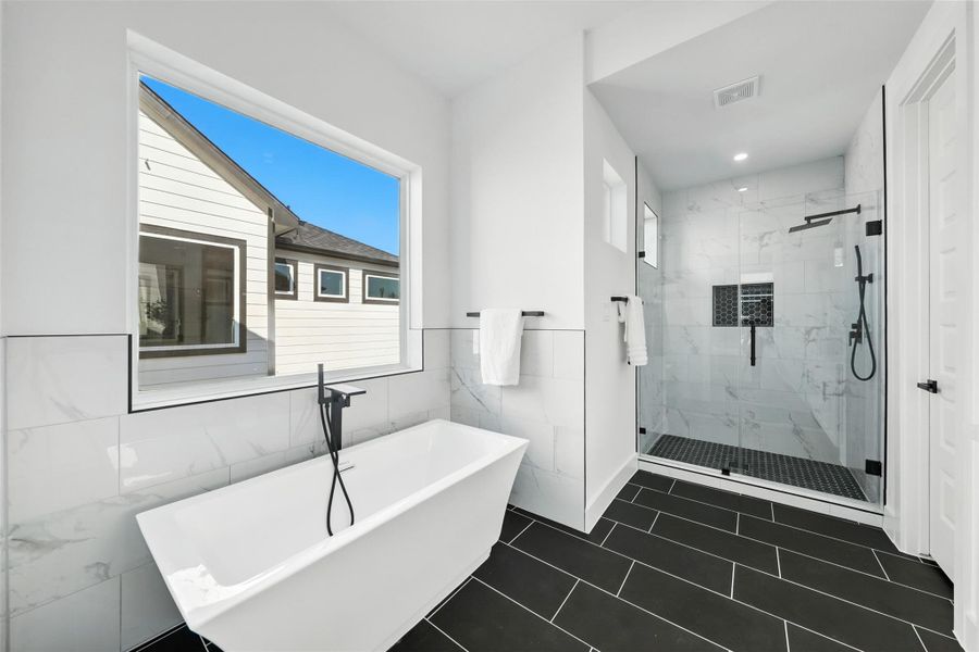 Spa-style primary bathroom featuring a freestanding soaking tub, frameless glass shower, and sleek tile finishes. Dual layout design with black hardware accents and large window for natural light—creating a clean, relaxing retreat. Spa-style primary bathroom featuring a freestanding soaking tub, frameless glass shower, and sleek tile finishes. Dual layout design with black hardware accents and large window for natural light—creating a clean, relaxing retreat.