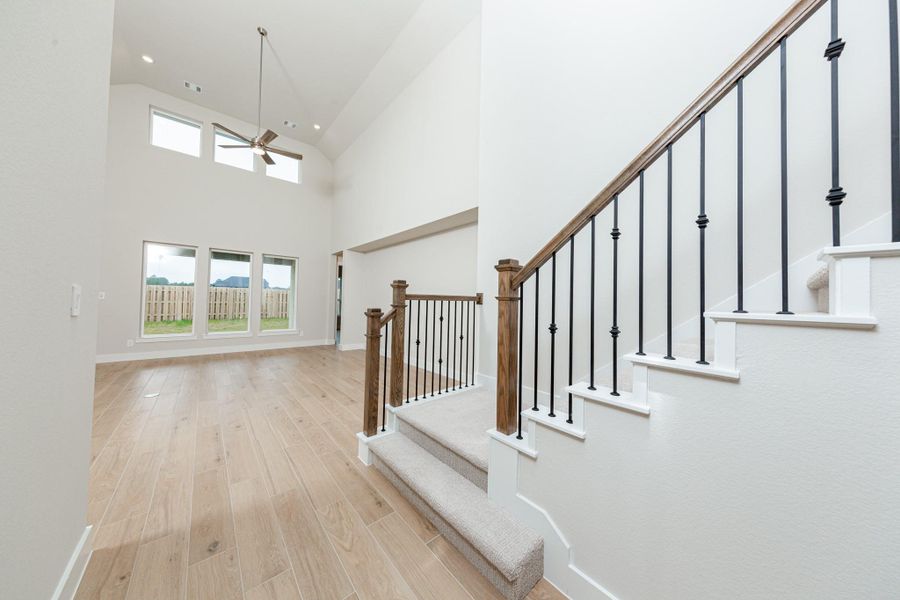 The staircase features iron rails and a rich wood handrail, contrasting elegantly with the light carpeted steps. The space is amplified by high ceilings and generous natural light pouring in from large windows, creating an airy and welcoming first impression.