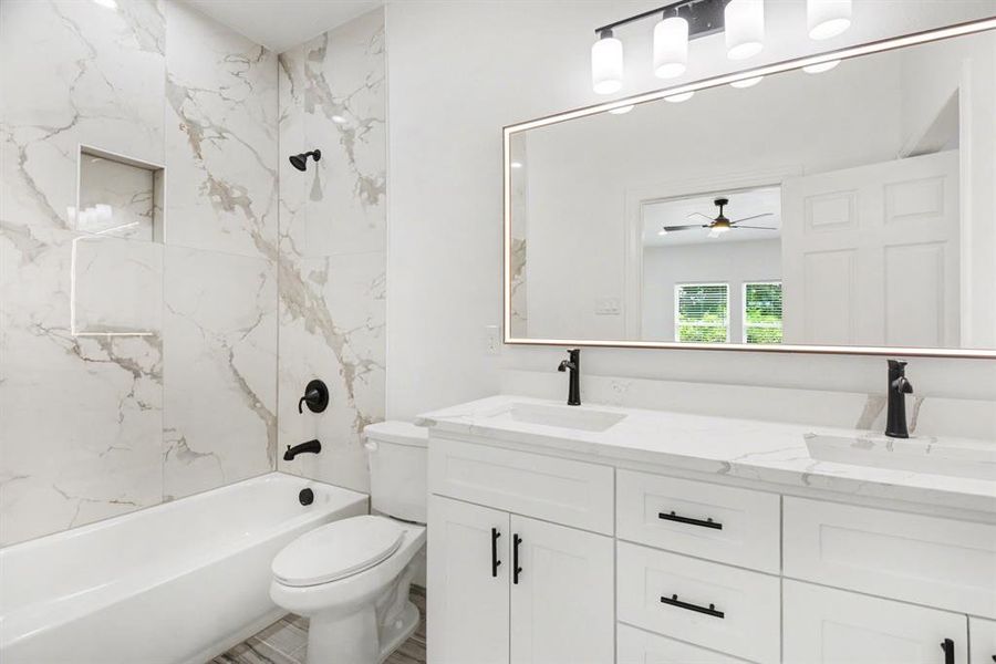 Bathroom featuring a white vanity with dual sinks, a large mirror with integrated lighting, a bathtub with a shower, and white marble-style wall tiles