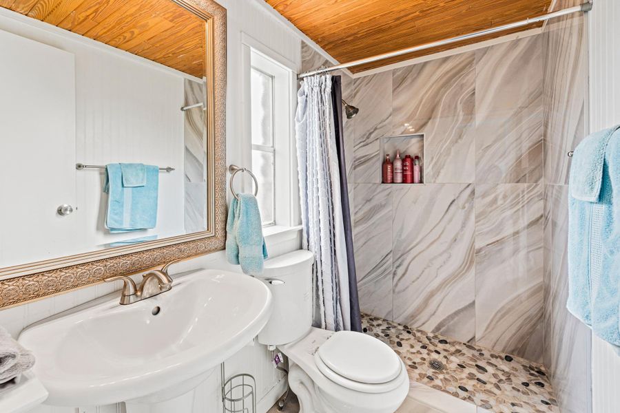 Bathroom featuring a wood-finish ceiling, a vanity with a white basin and brushed nickel faucet, a framed mirror, a toilet, and a shower with stone-look tile walls and a pebble floor