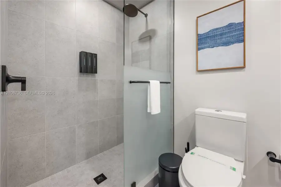 Walk-in shower featuring large-format grey tile, a glass partition with a frosted privacy panel, and matte black fixtures including a wall-mounted shower head and faucet Walk-in shower featuring large-format grey tile, a glass partition with a frosted privacy panel, and matte black fixtures including a wall-mounted shower head and faucet