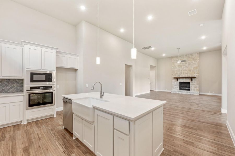 Open kitchen with white island, farmhouse sink, stainless appliances, and stone fireplace in adjoining living room