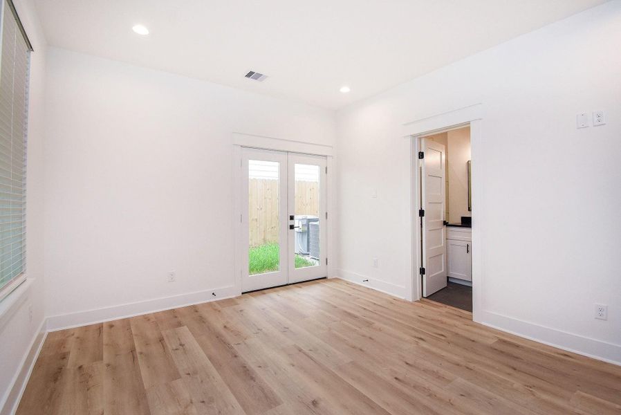This bright room features light wood flooring, white walls, and recessed lighting. It includes French doors leading to a fenced backyard and an adjacent bathroom with modern fixtures. Ideal for a cozy bedroom or office space.