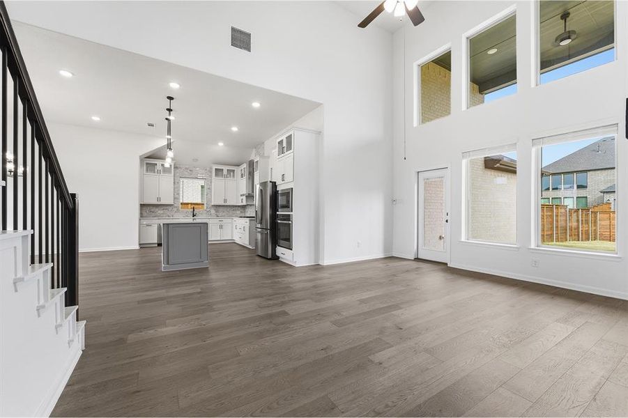 Open-concept living area featuring high ceilings, wood-finish flooring, a modern kitchen with a gray island, and abundant natural light from multiple large windows