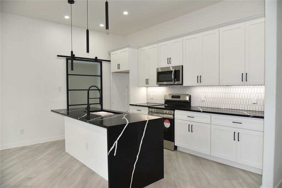 Kitchen with appliances with stainless steel finishes, a kitchen island with sink, a barn door, white cabinetry, and hanging light fixtures