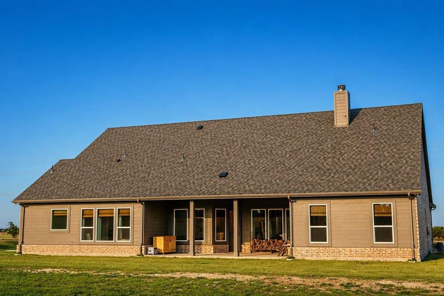 Rear exterior featuring a covered patio, multiple windows, a shingled roof, a chimney, and a brick base