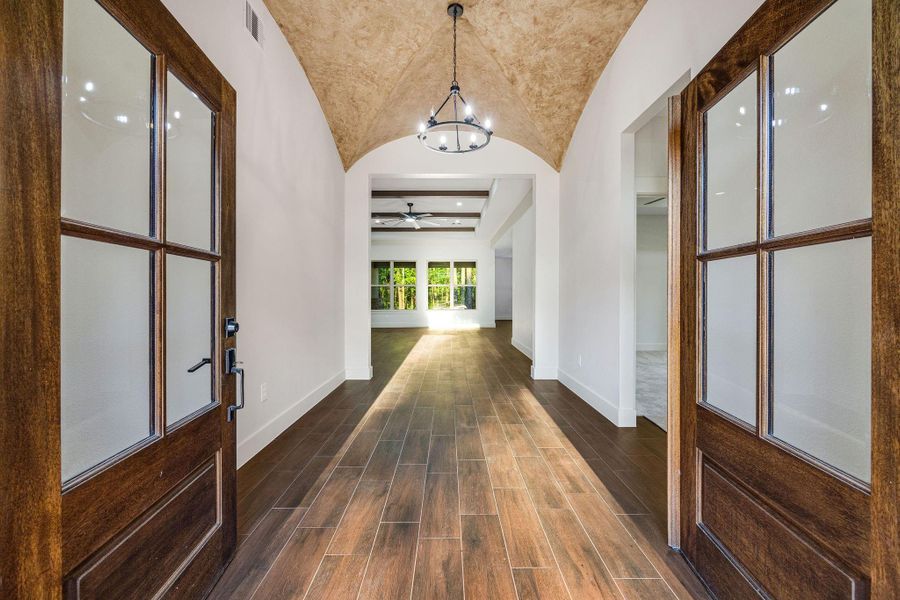 This entryway features elegant wooden double doors leading to a spacious hallway with rich wood flooring and a vaulted ceiling. A stylish chandelier adds charm, while large windows at the end offer natural light and a view of the greenery outside.
