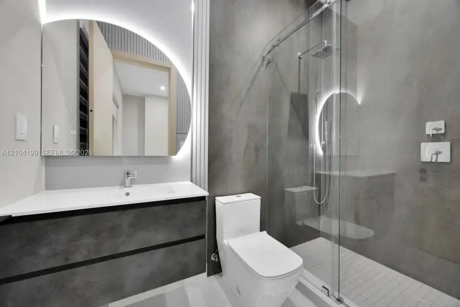 Bathroom featuring a floating vanity with a white countertop, an arched LED-backlit mirror, large-format grey tile walling, and a walk-in shower with a glass enclosure
