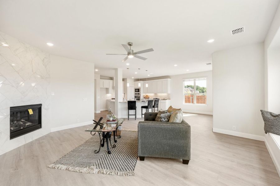 Open-concept living room with marble tile fireplace, hardwood floors, and ceiling fan, open to white kitchen with island seating.