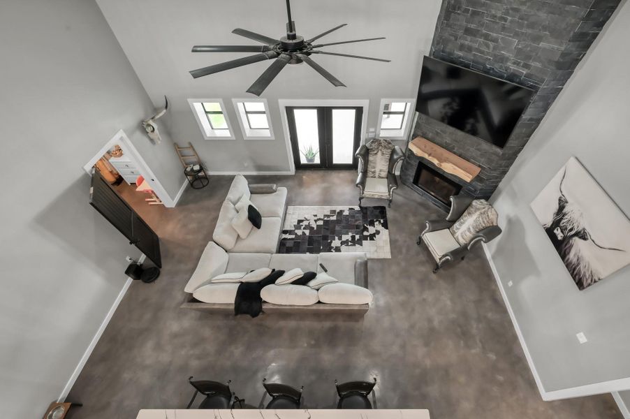 Another view of this spacious family room featuring a modern open layout with high ceilings, a large ceiling fan, and a cozy fireplace. The room is well-lit with natural light from the windows and glass doors.