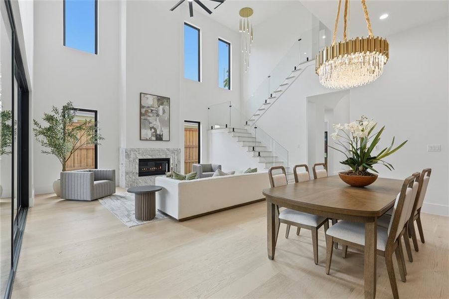 Open-concept living space featuring a two-story ceiling, light wood-finish flooring, a contemporary fireplace with a stone surround, an elegant staircase with glass railings, and a prominent chandelier Open-concept living space featuring a two-story ceiling, light wood-finish flooring, a contemporary fireplace with a stone surround, an elegant staircase with glass railings, and a prominent chandelier