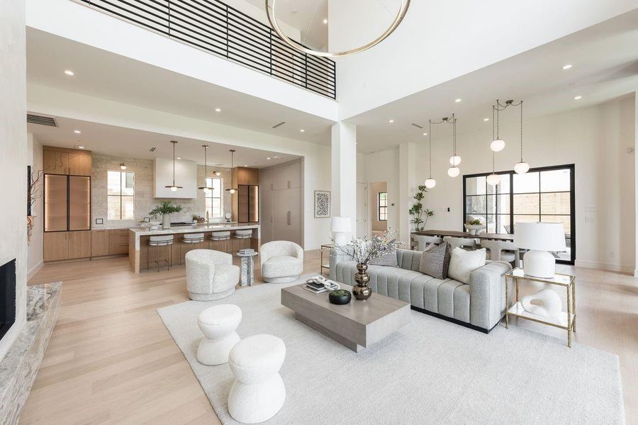 From the kitchen, sightlines extend effortlessly into a dramatic two-story living space where towering windows allow natural light to pour in. A sleek linear fireplace anchors the room, while floating shelves and warm textures create an atmosphere that feels both curated and comfortably lived-in.
