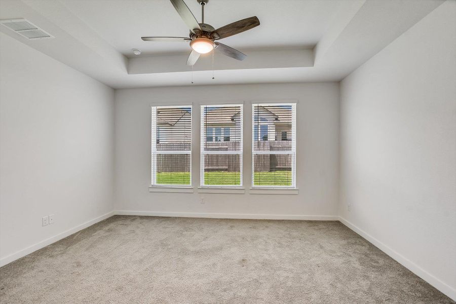 Spacious Primary bedroom featuring a tray ceiling, ceiling fan with integrated lighting, three large windows, textured carpeting, and white baseboards