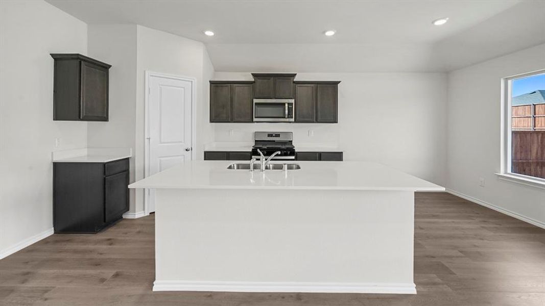 Kitchen featuring appliances with stainless steel finishes, a kitchen island with sink, light stone countertops, recessed lighting, and dark brown cabinets