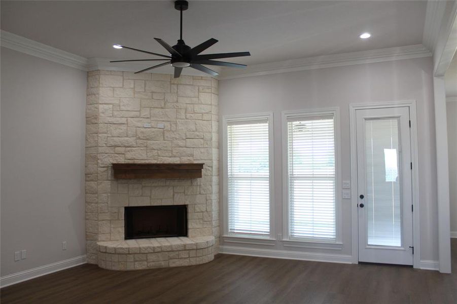Unfurnished living room with ceiling fan, crown molding, dark wood finished floors, a stone fireplace, and plenty of natural light Unfurnished living room with ceiling fan, crown molding, dark wood finished floors, a stone fireplace, and plenty of natural light