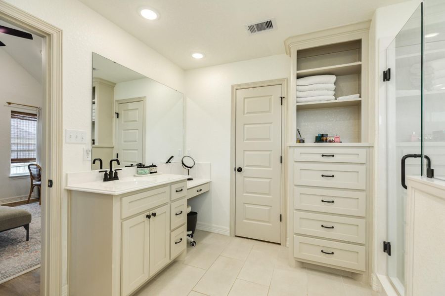This photo showcases a bright, modern bathroom with a large mirror above a vanity. It features ample storage with built-in cabinets and drawers, sleek black fixtures, and a tile floor. The door leads to a large walk-in closet, enhancing accessibility and flow.