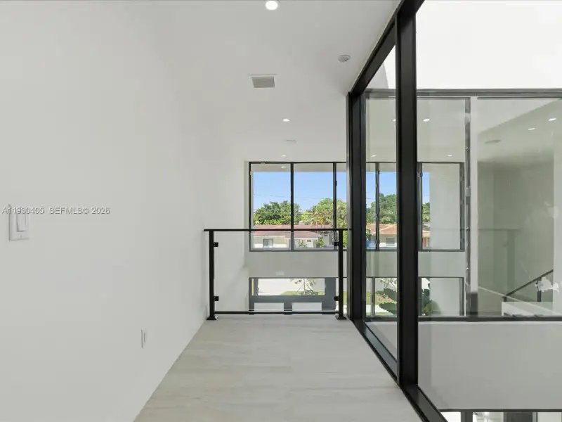 Light-filled interior featuring floor-to-ceiling black-frame windows, recessed lighting, and a glass-panel railing with black metal hardware, finished with neutral large-format tile flooring Light-filled interior featuring floor-to-ceiling black-frame windows, recessed lighting, and a glass-panel railing with black metal hardware, finished with neutral large-format tile flooring