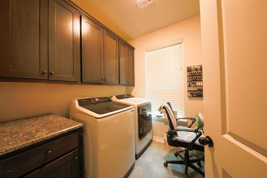 This photo showcases a cozy laundry room with dark wood cabinets, a granite countertop, and modern washer and dryer units. A window provides natural light, and there's a small desk with a chair for added functionality.