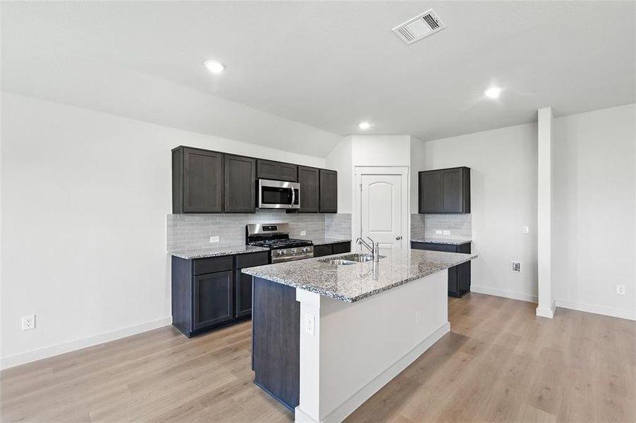 Kitchen featuring stainless steel appliances, light stone countertops, light wood finished floors, backsplash, and a kitchen island with sink