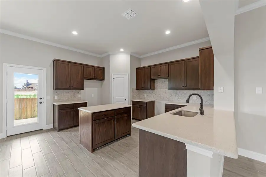 Kitchen featuring decorative backsplash, ornamental molding, dark brown cabinets, a peninsula, and light stone countertops