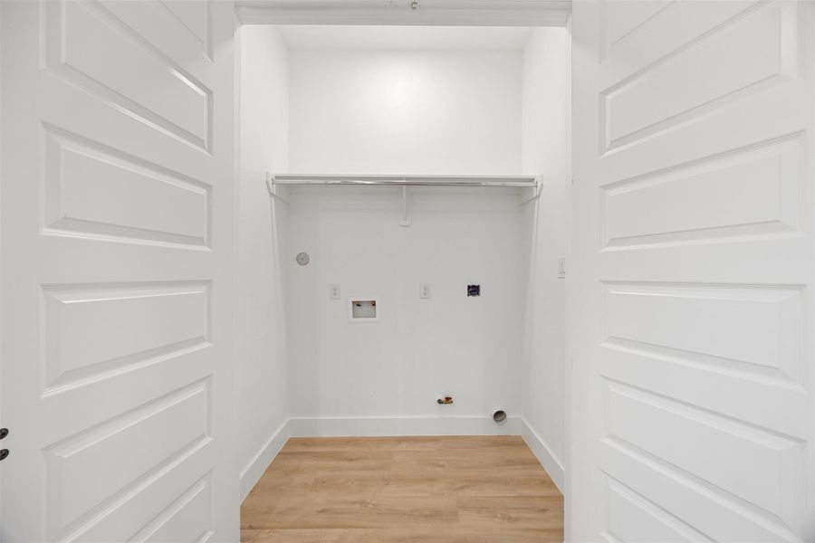 This photo shows a bright, clean laundry room with white walls and wood-like flooring. It features hookups for a washer and dryer, a shelf for storage, and double doors for easy access.
