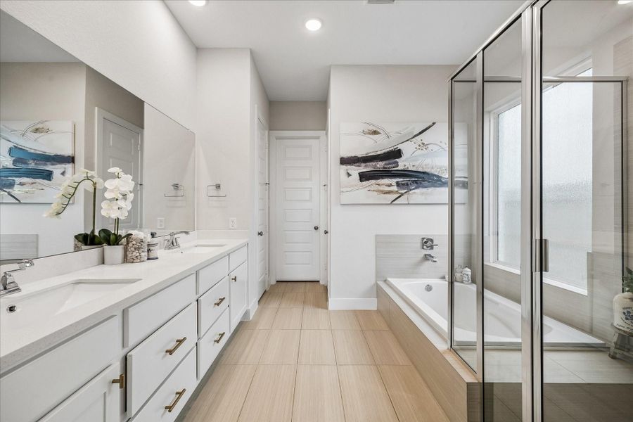 The spa-inspired primary bathroom features wood-look tile flooring, dual vanities with white cabinetry and gold hardware, a large glass-enclosed shower, and a relaxing soaking tub.